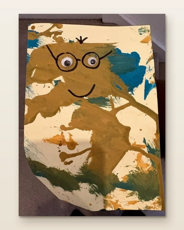 A phone photo of a child's painting on a carpet: a brown paint face on cream card, with googly eyes and marker glasses.