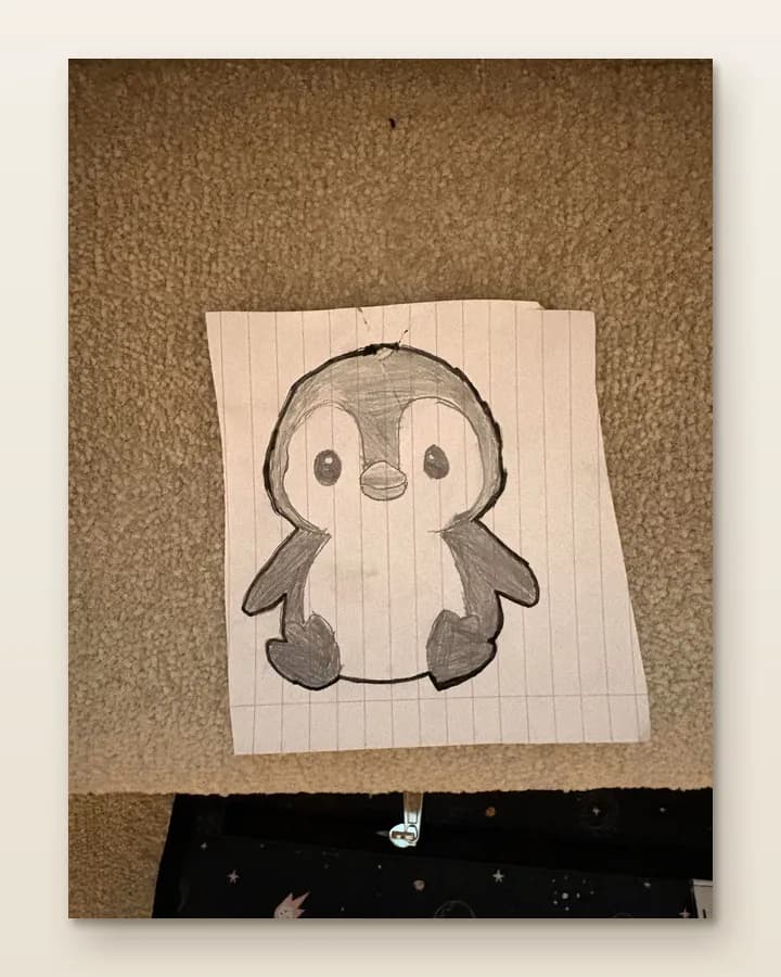A phone photo of a child's pencil drawing of a penguin, shaded grey with a pale belly, on a sheet of lined paper torn from a pad, lying on a carpet under a lamp.